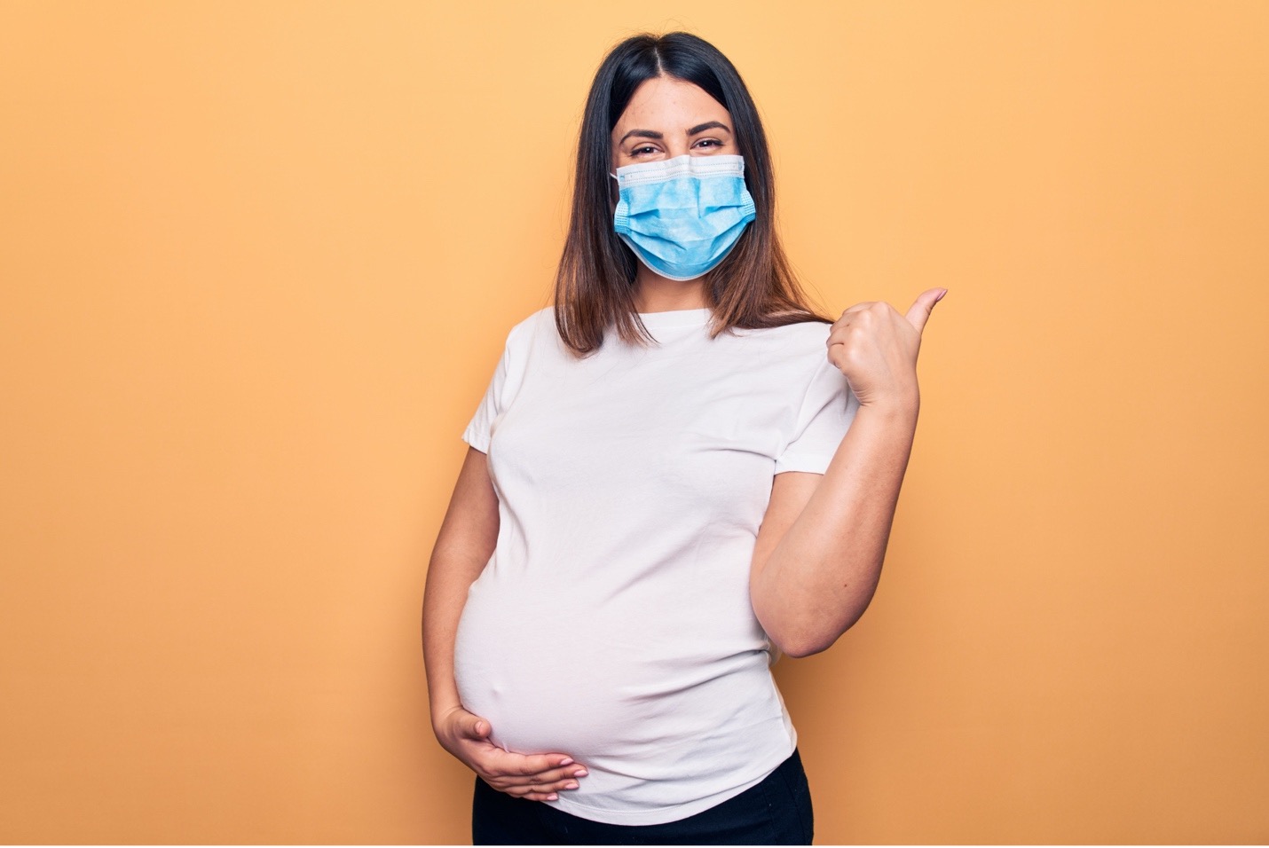 A pregnant individual wearing a medical mask gives a thumbs up