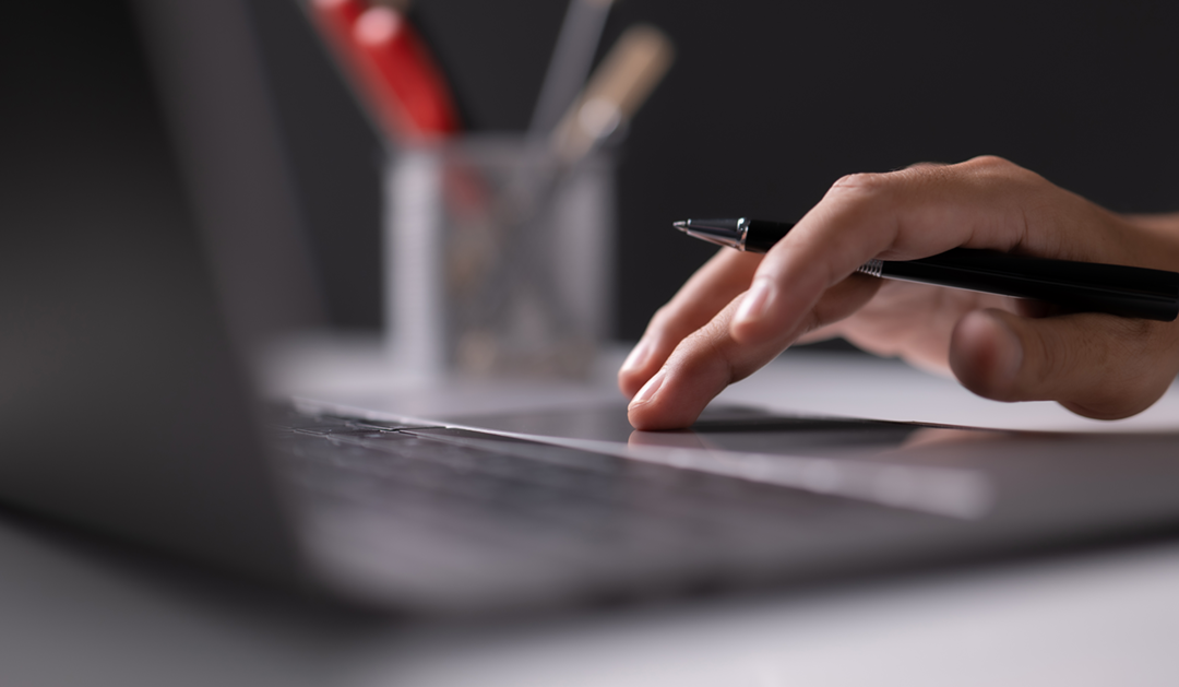 A person holds a pen in their hand while typing on a laptop